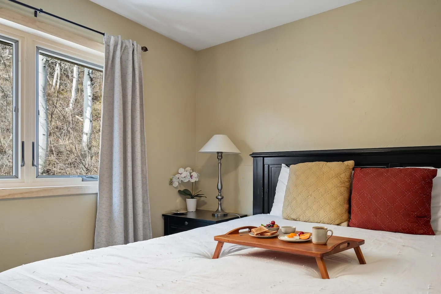 Bedroom with a wooden breakfast tray on a white-made bed next to a window showing outdoor trees.