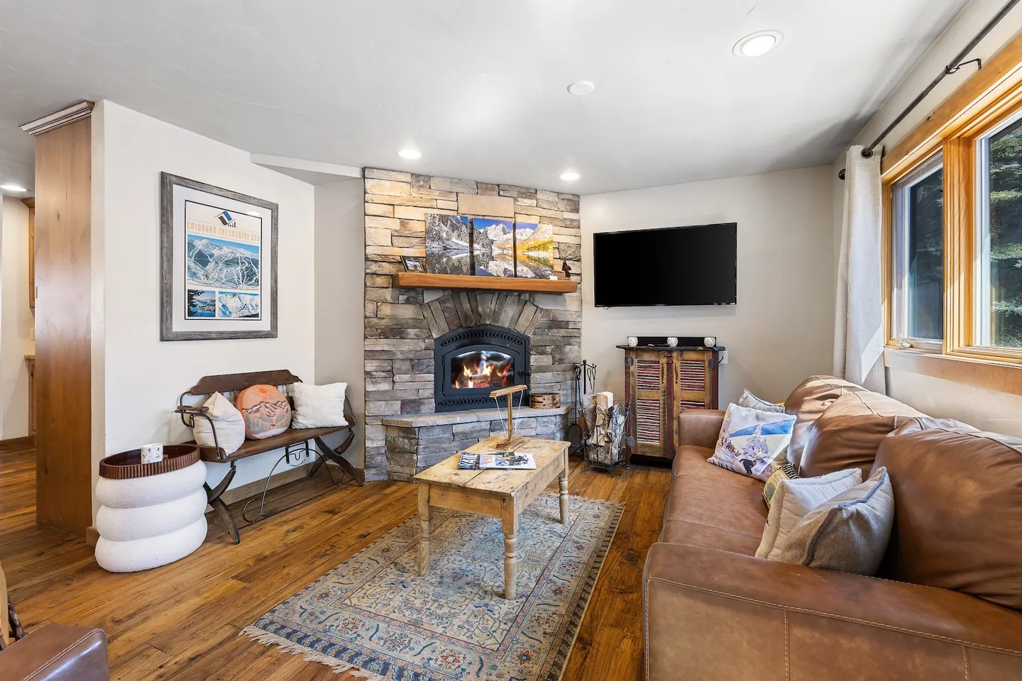 Living room with a stone corner fireplace, leather sofa, wood coffee table, patterned area rug, and wall-mounted television.