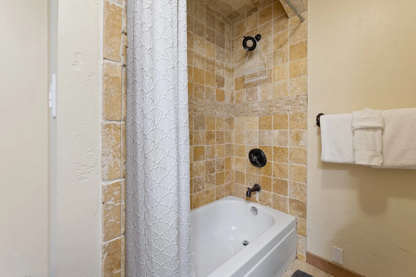 Bathroom bathtub and shower combination surrounded by beige stone-look tile with dark hardware and a textured white shower curtain.