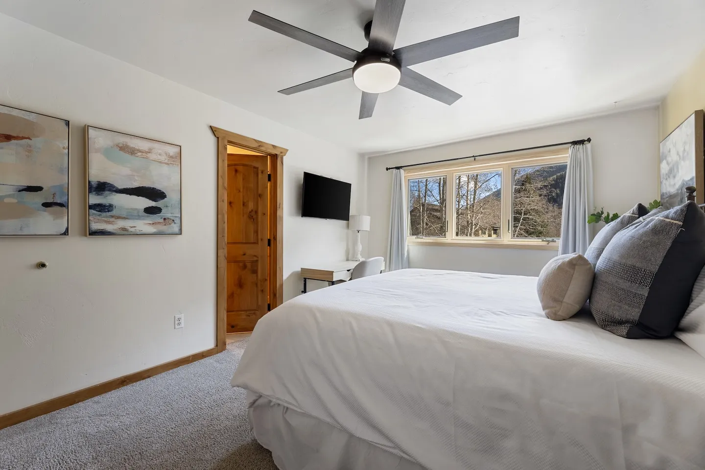 Bedroom with a large bed dressed in white linens, carpeted floor, ceiling fan, wall-mounted television, and a window with mountain views.
