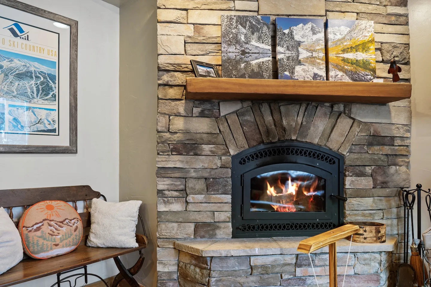 Stone fireplace with a lit fire, a wooden mantel displaying landscape photos, and an adjacent bench with cushions.