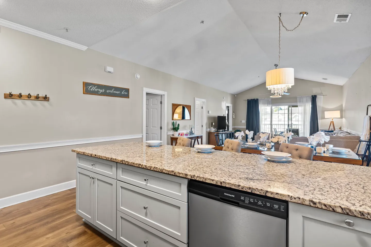 Open-concept living space viewed from a kitchen island with granite countertops, showing a dining area and living room with vaulted ceilings.