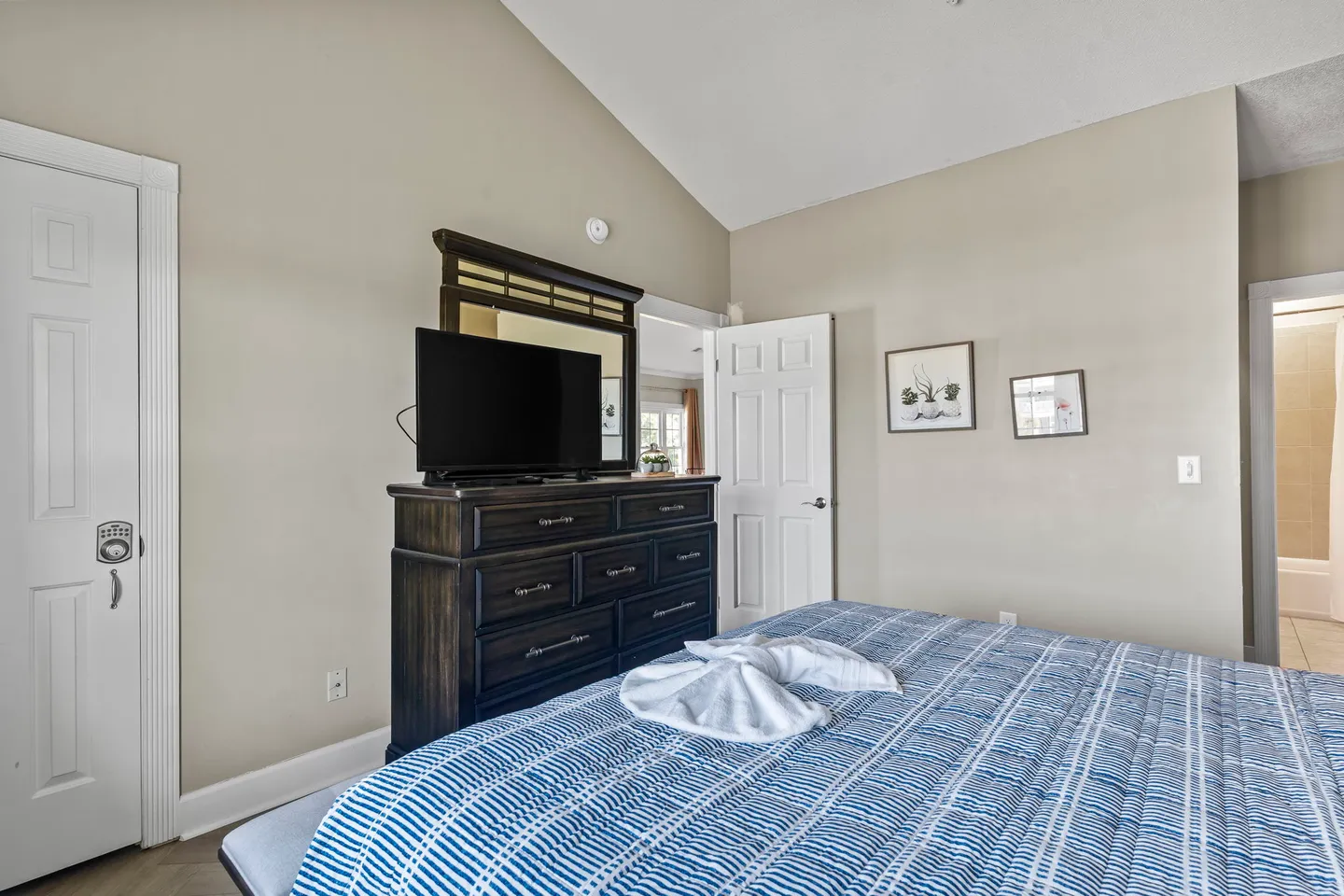 Bedroom featuring a bed with a blue and white patterned spread, a dark wood dresser with a mounted mirror and flat-screen TV, and high angled ceilings.