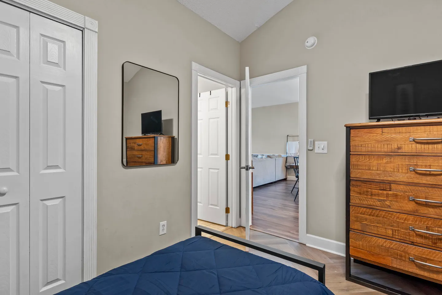 Bedroom featuring a wooden dresser with a TV, a wall mirror, a bed with a blue quilt, and a view into an adjacent living area.