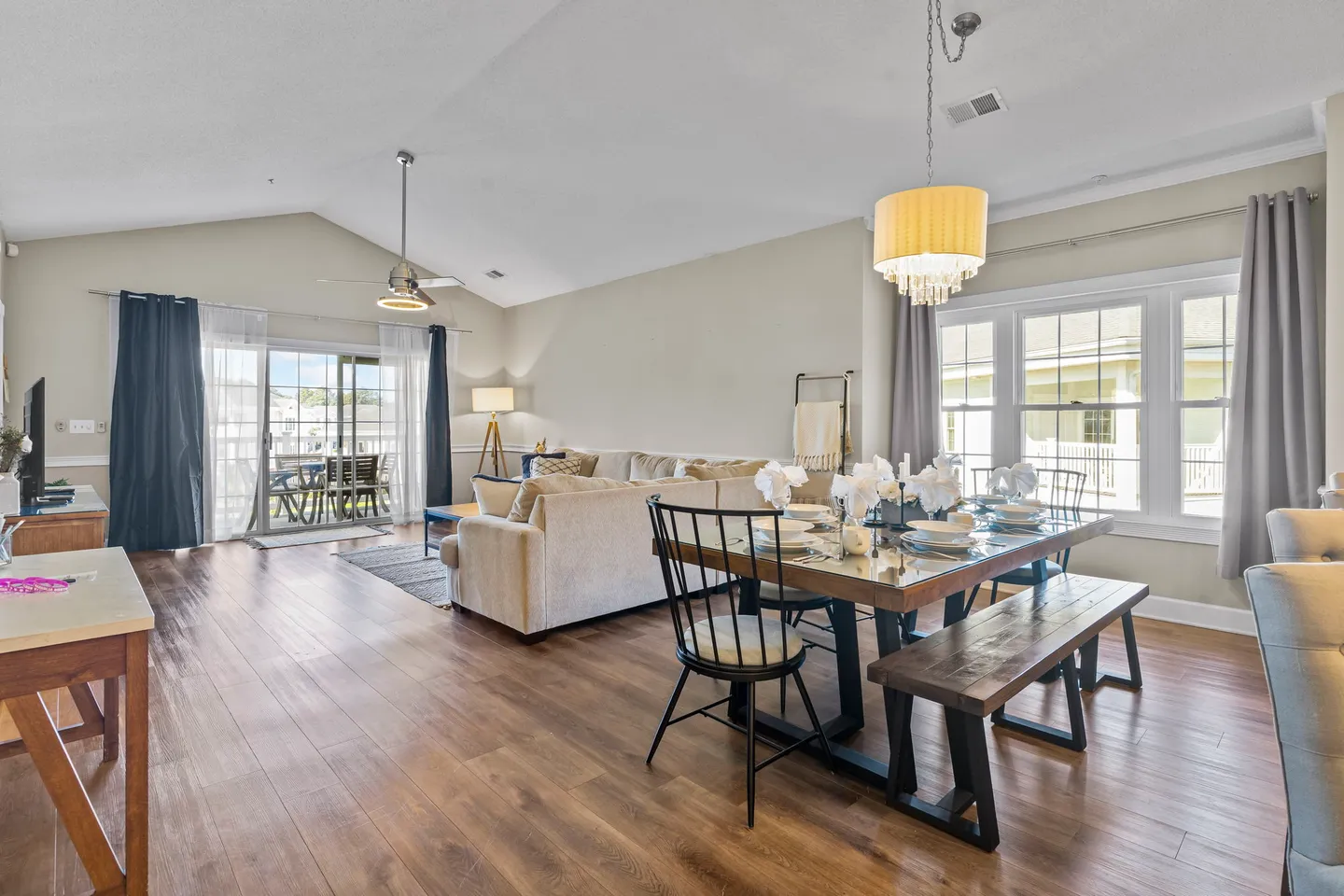 Open-concept living and dining area with vaulted ceiling, wood-look flooring, dining table with bench, and sliding glass doors leading to a balcony.