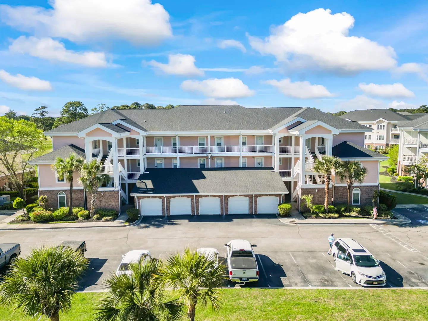 Elevated view of a three-story multi-family residential building with ground-level garages, palm trees, and an open parking lot.