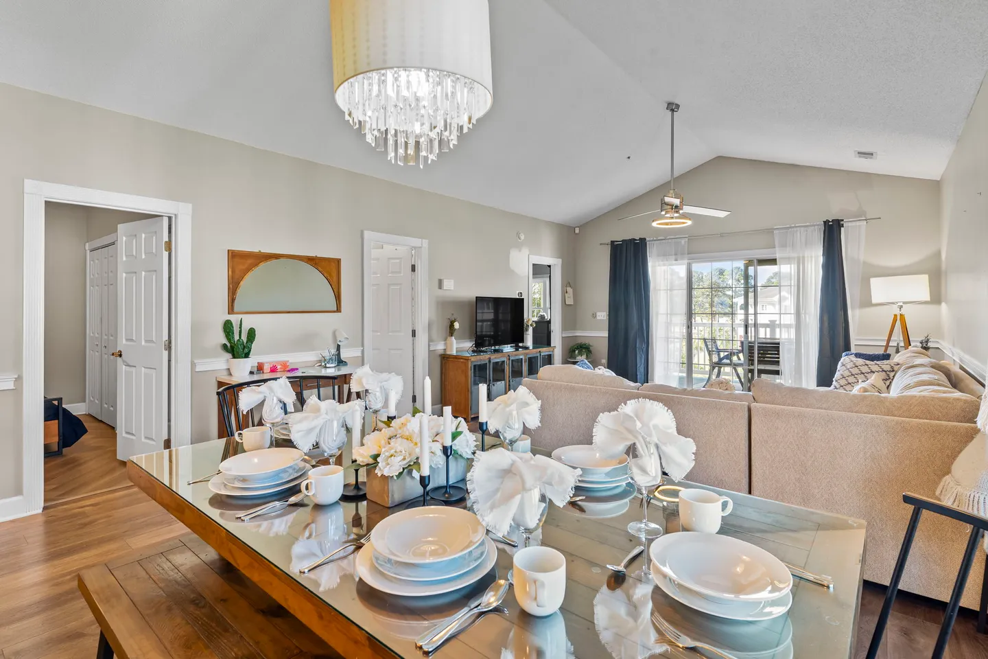 Dining area with a set glass-top table under a crystal-accented chandelier, open to an adjoining living room with vaulted ceiling and sliding glass doors.