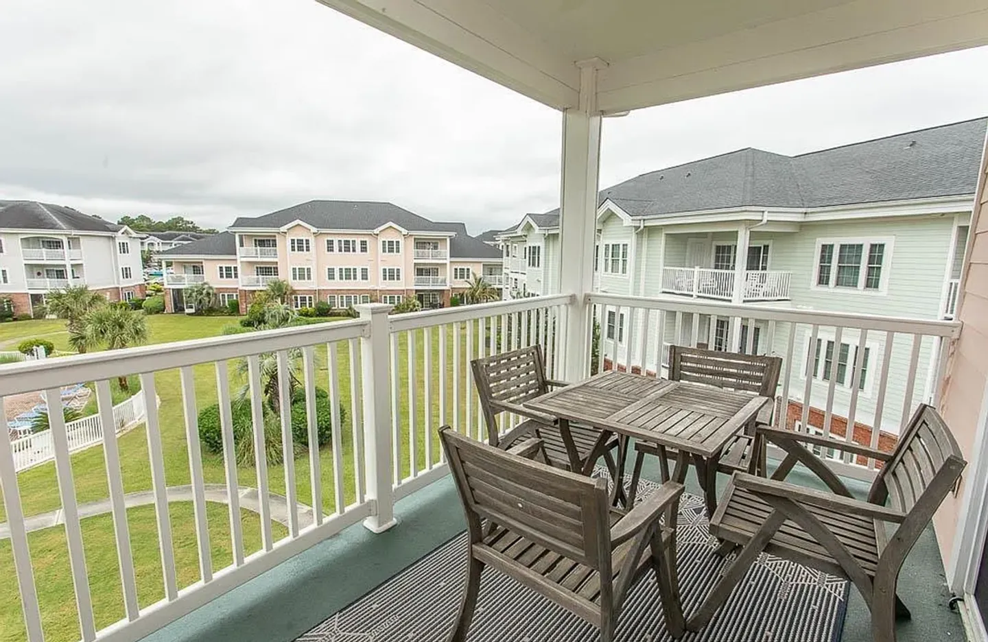 Covered outdoor balcony with a wooden table, four wooden chairs, and a patterned rug overlooking a grassy courtyard and neighboring apartment buildings.