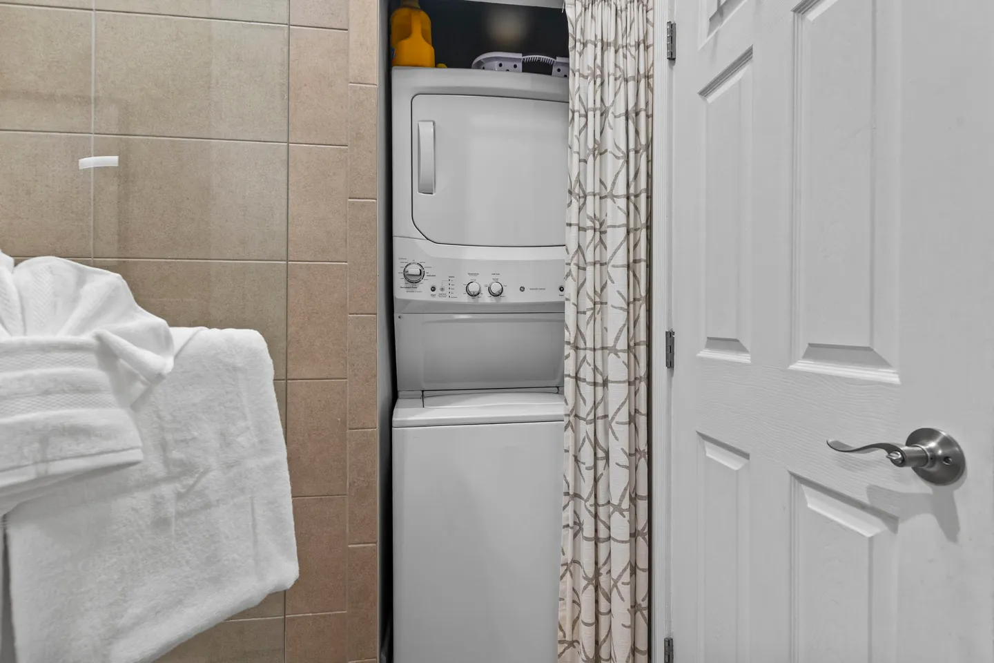 A stacked white washing machine and dryer unit tucked into a closet space next to a tiled wall and open white door.