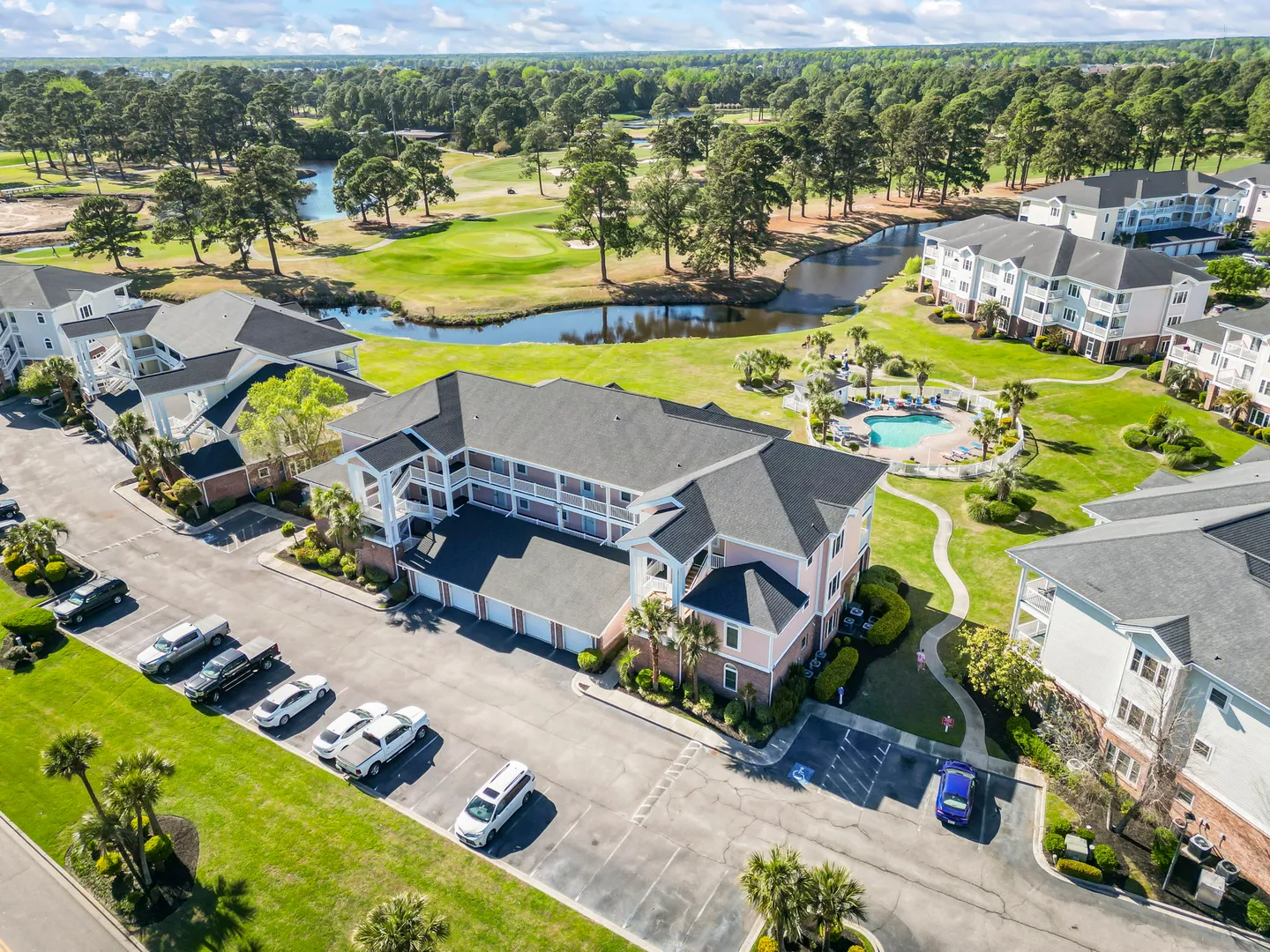 Aerial view of a multi-story condo complex with parking lot, swimming pool, pond, and adjacent golf course.