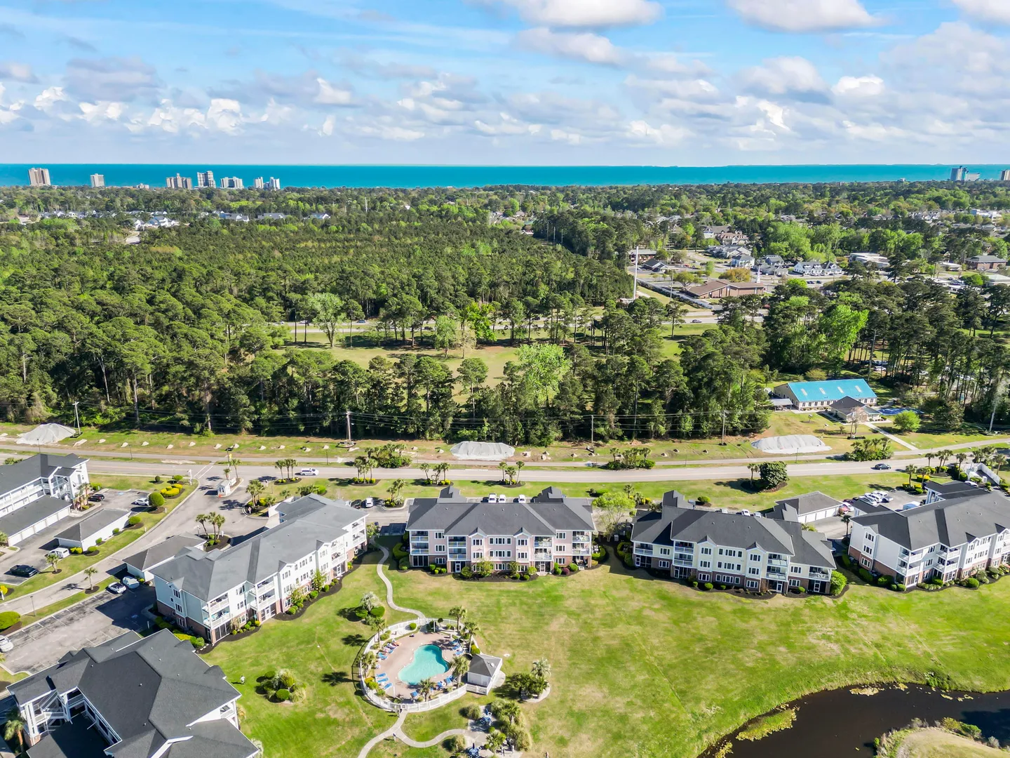 Aerial view of a multi-building residential complex with a central outdoor pool, extensive green lawns, surrounding woodland, and the ocean along the horizon.