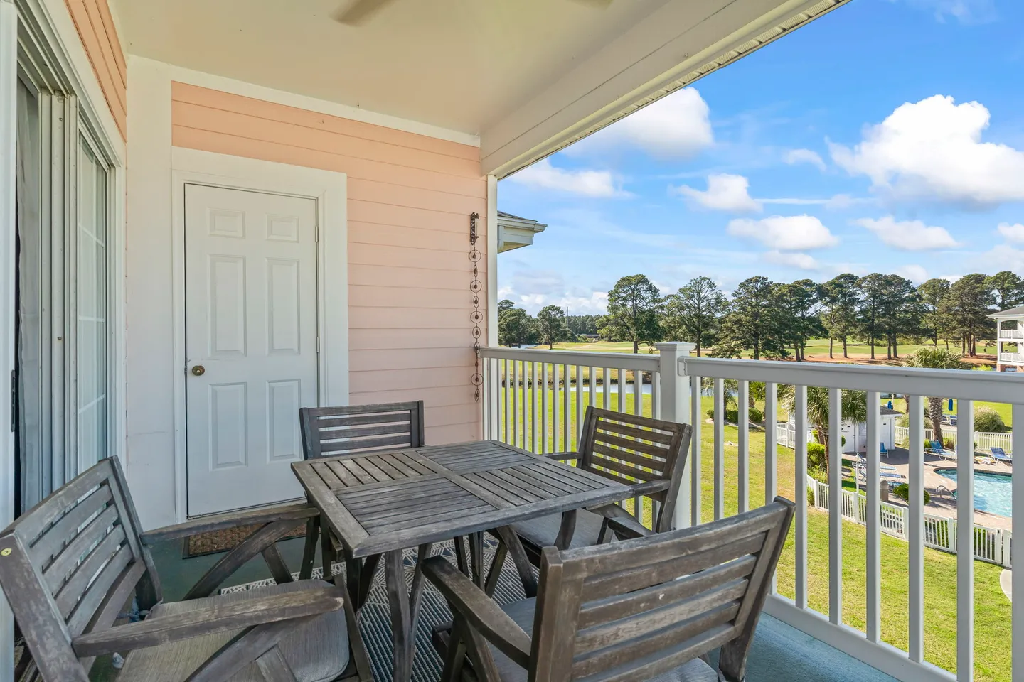 Balcony with a wooden outdoor dining table and four chairs overlooking a golf course and pool area.