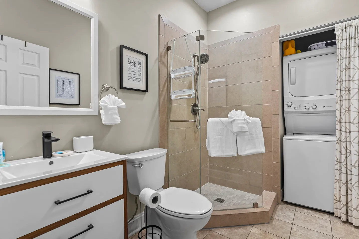 Bathroom with a modern vanity, toilet, glass-enclosed corner walk-in shower, and a stacked washer and dryer unit in a closet.