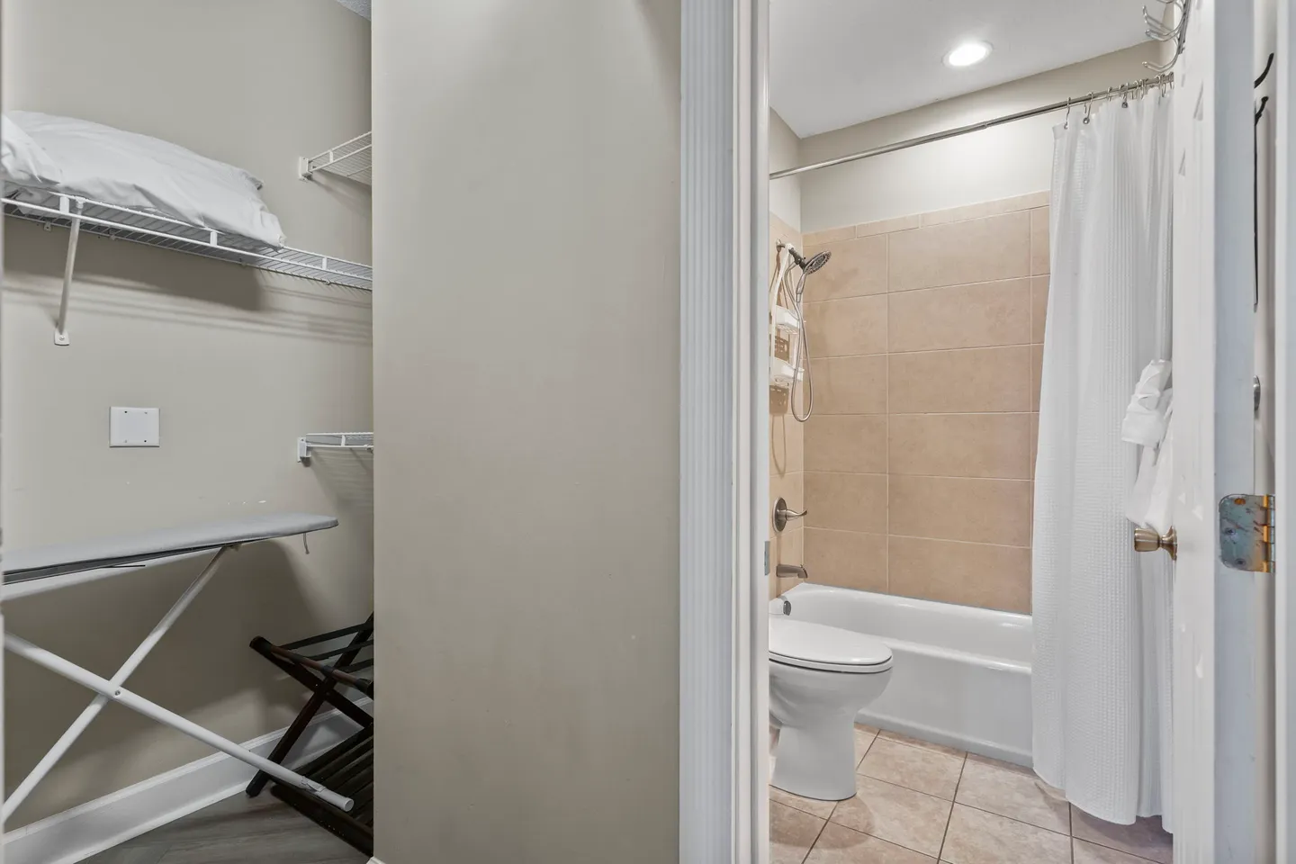 A view through an open doorway showing a walk-in closet with an ironing board on the left and a bathroom with a toilet and shower-tub combo on the right.
