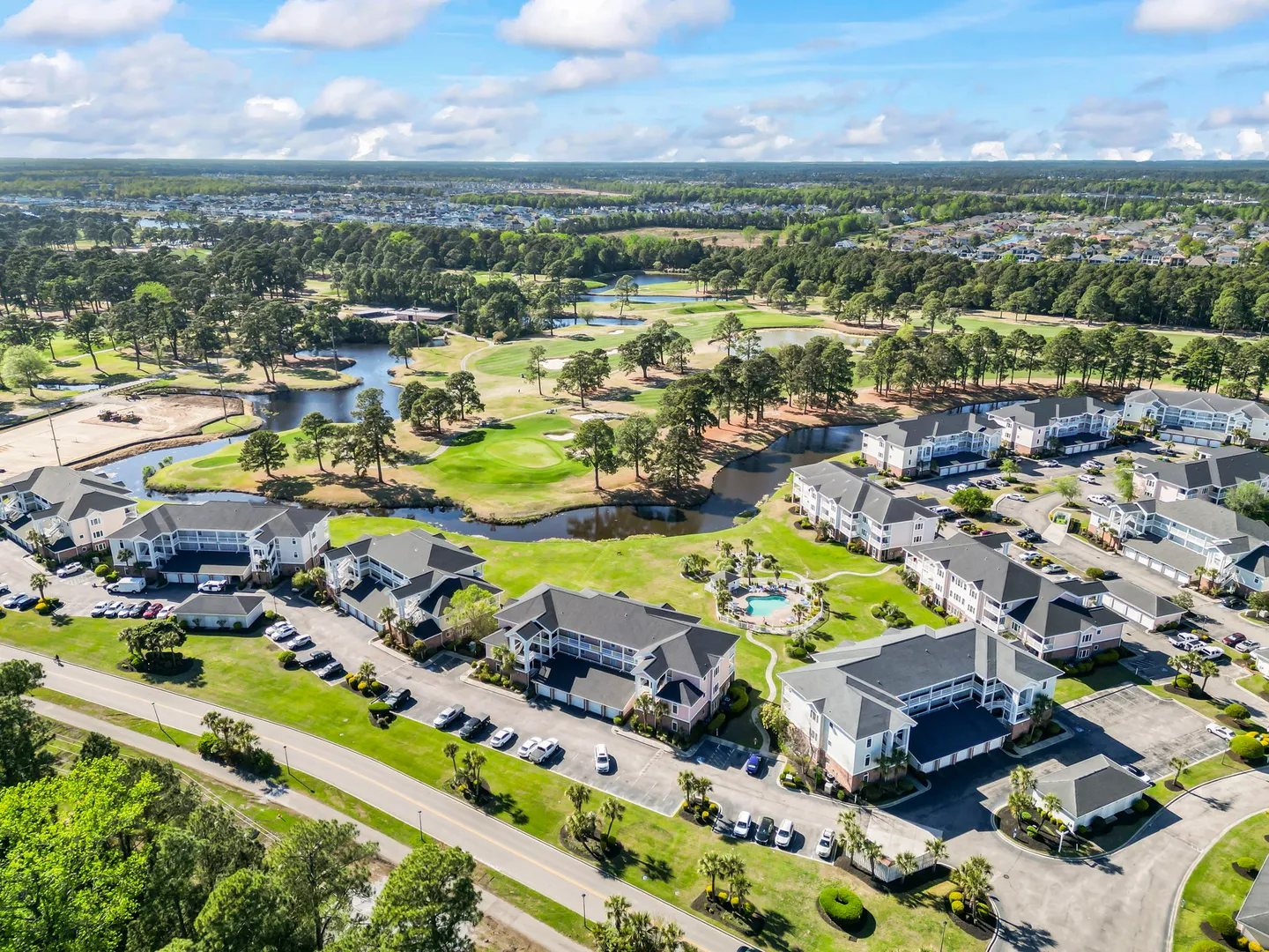 Aerial view of a condominium complex next to a golf course with ponds, surrounded by trees under a blue sky.