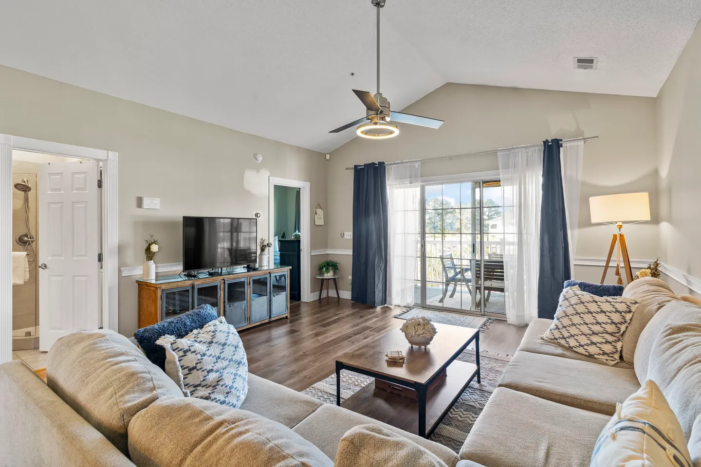 Living room with a large sectional sofa, coffee table, television on a credenza, vaulted ceiling with fan, and sliding glass doors leading to a balcony.