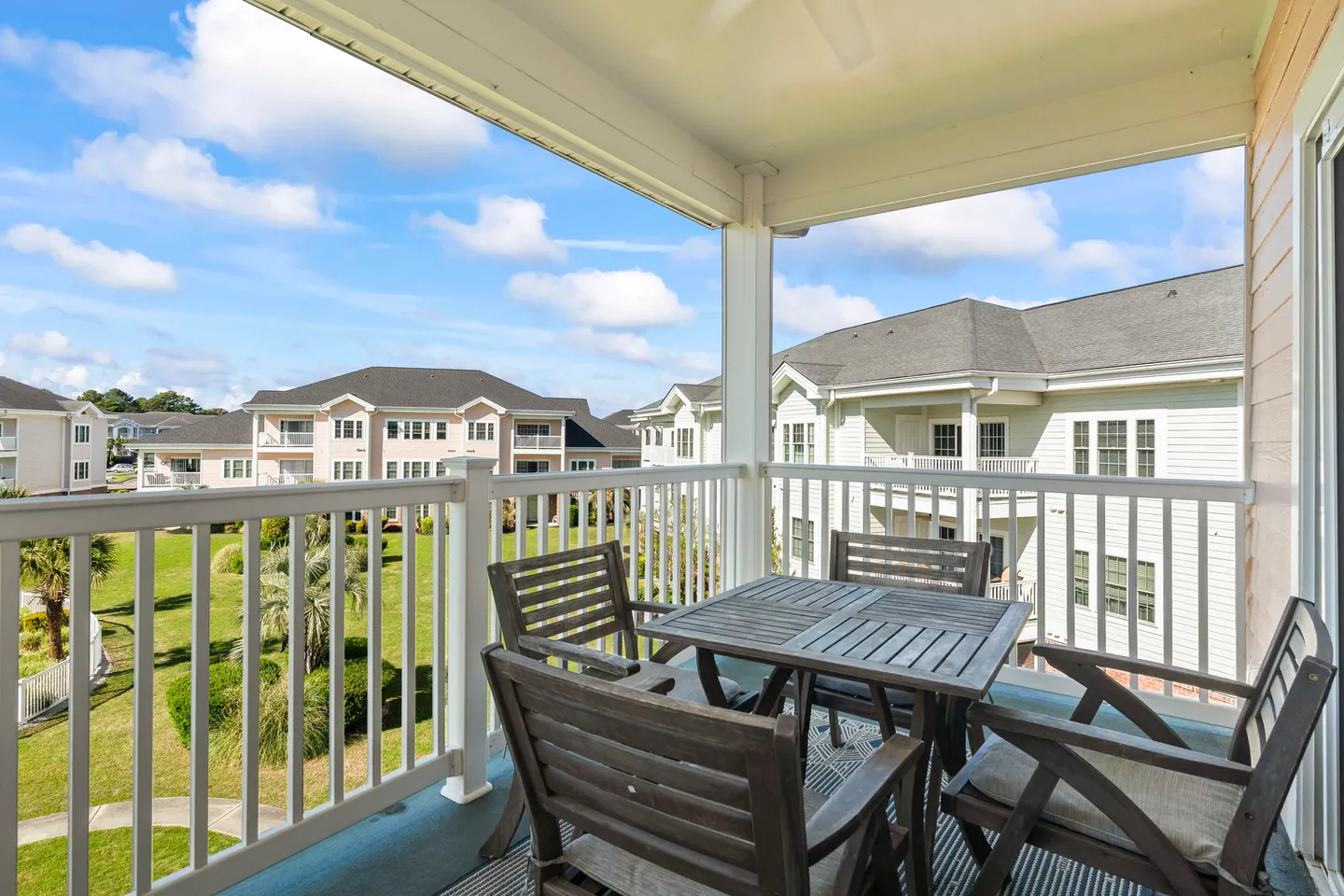 Covered outdoor balcony with a slatted table and four wooden armchairs overlooking residential buildings and a green lawn under a partly cloudy blue sky.