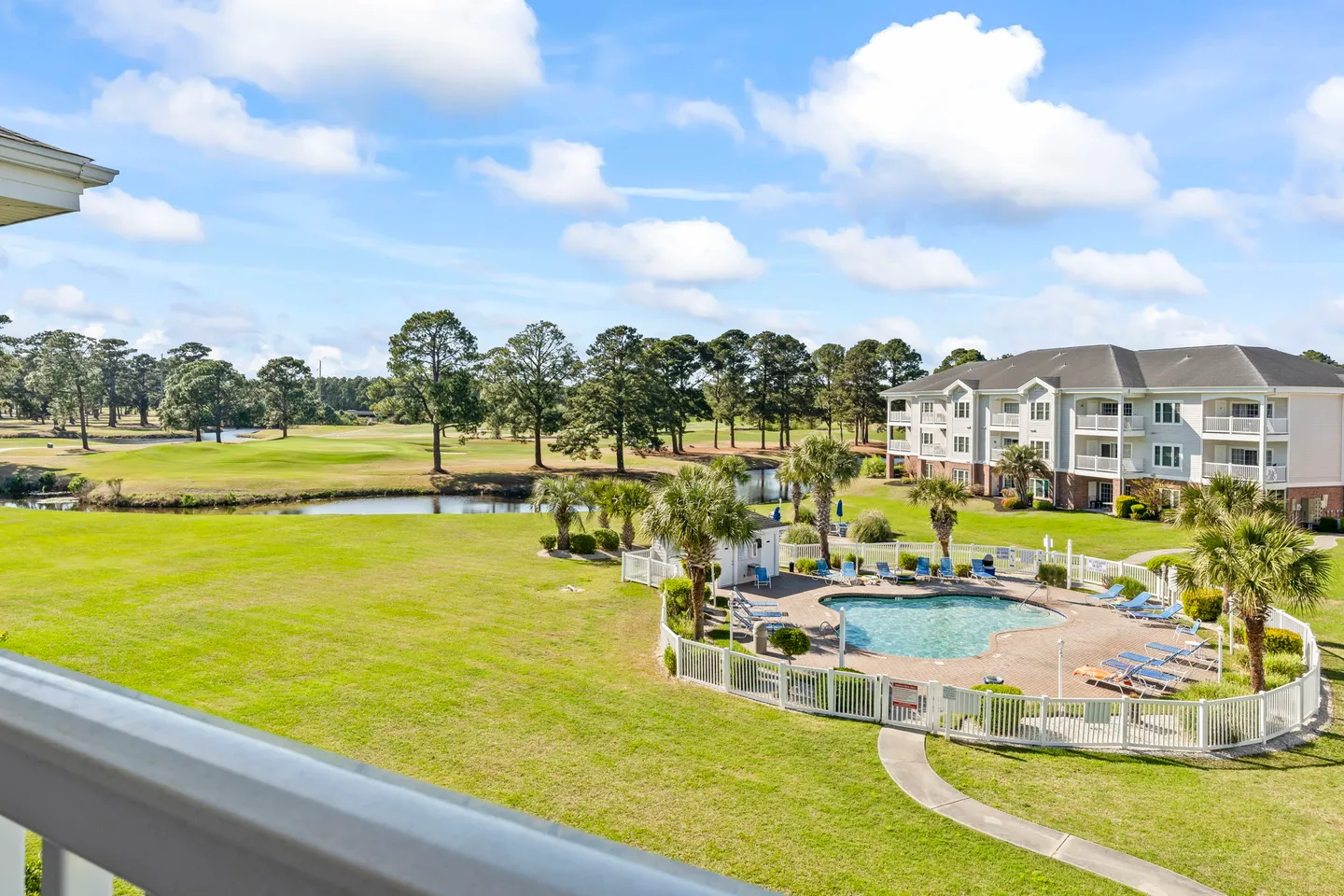 Balcony view overlooking a fenced community swimming pool, pond, green golf course, and apartment buildings under a partly cloudy sky.