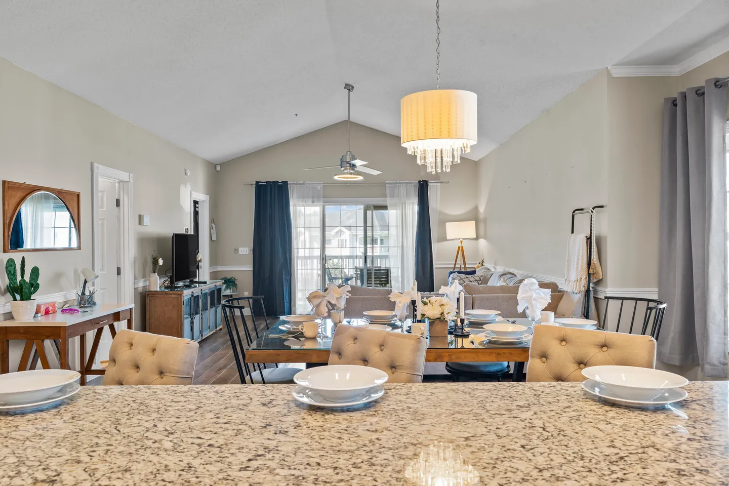 Open-concept dining and living area viewed over a granite kitchen counter toward a set dining table and sliding glass balcony doors.