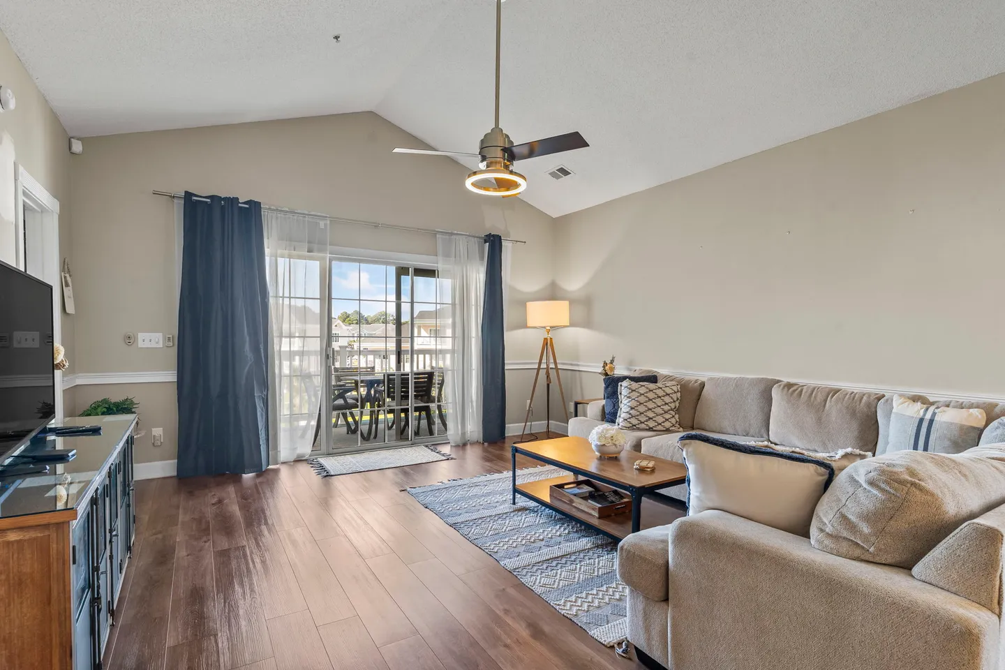 Living room with vaulted ceiling, ceiling fan, sectional sofa, wood-look flooring, and sliding glass doors leading to an outdoor balcony.
