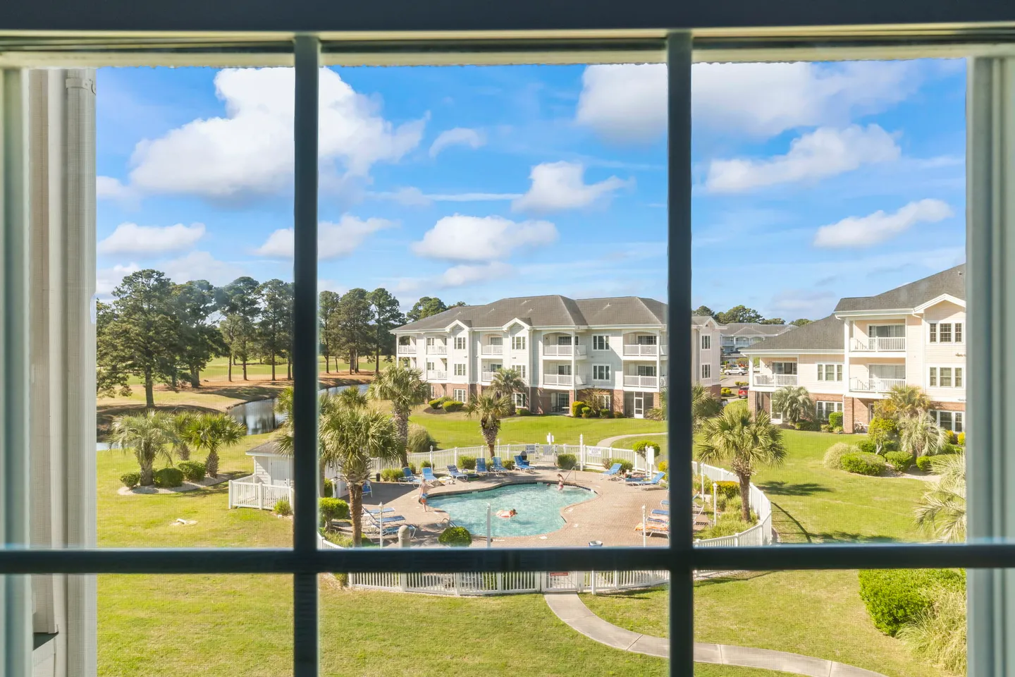 View through a gridded window overlooking an outdoor swimming pool, apartment buildings, palm trees, and green lawns under a blue sky.