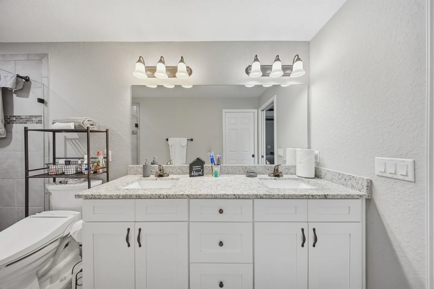 Bathroom with a double vanity, large frameless mirror, two vanity light fixtures, and a toilet next to a shelving unit.