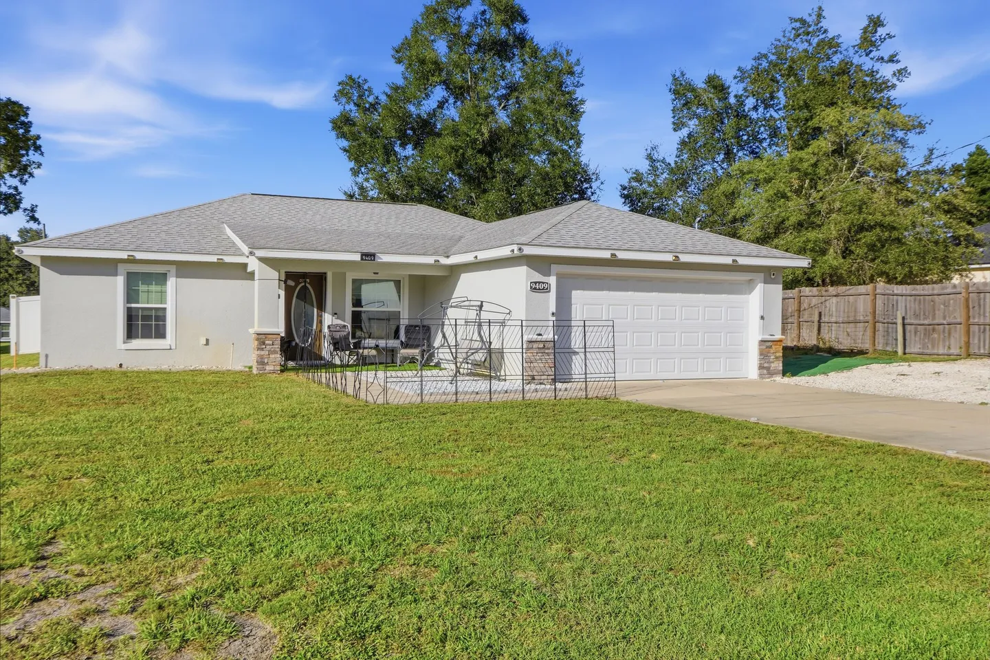 Exterior front view of a single-story home with a grass lawn, two-car garage, and a fenced front patio area under a bright blue sky.