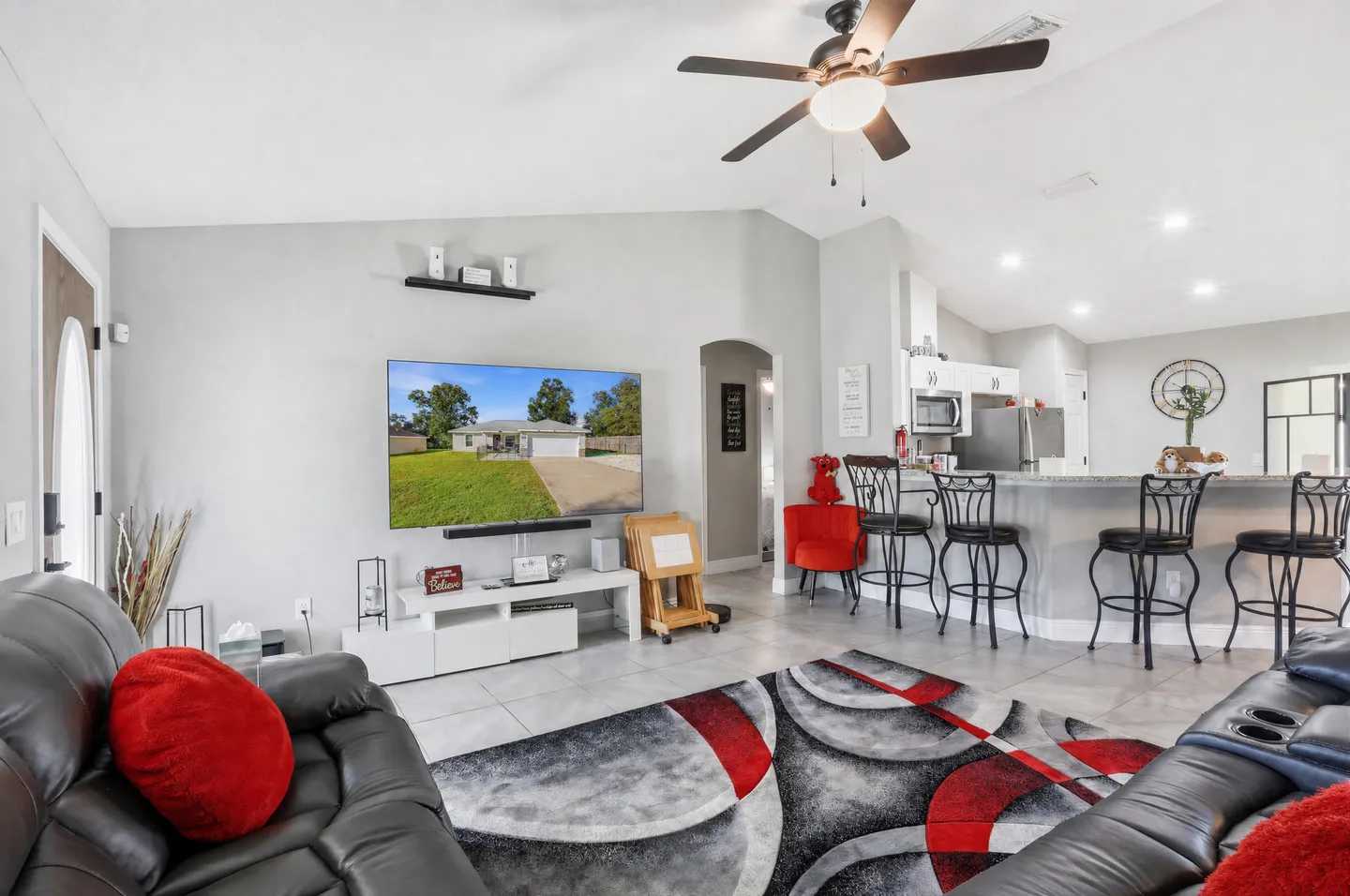 Living room with black leather recliners, a wall-mounted TV, patterned area rug, and an adjacent kitchen bar with stools.