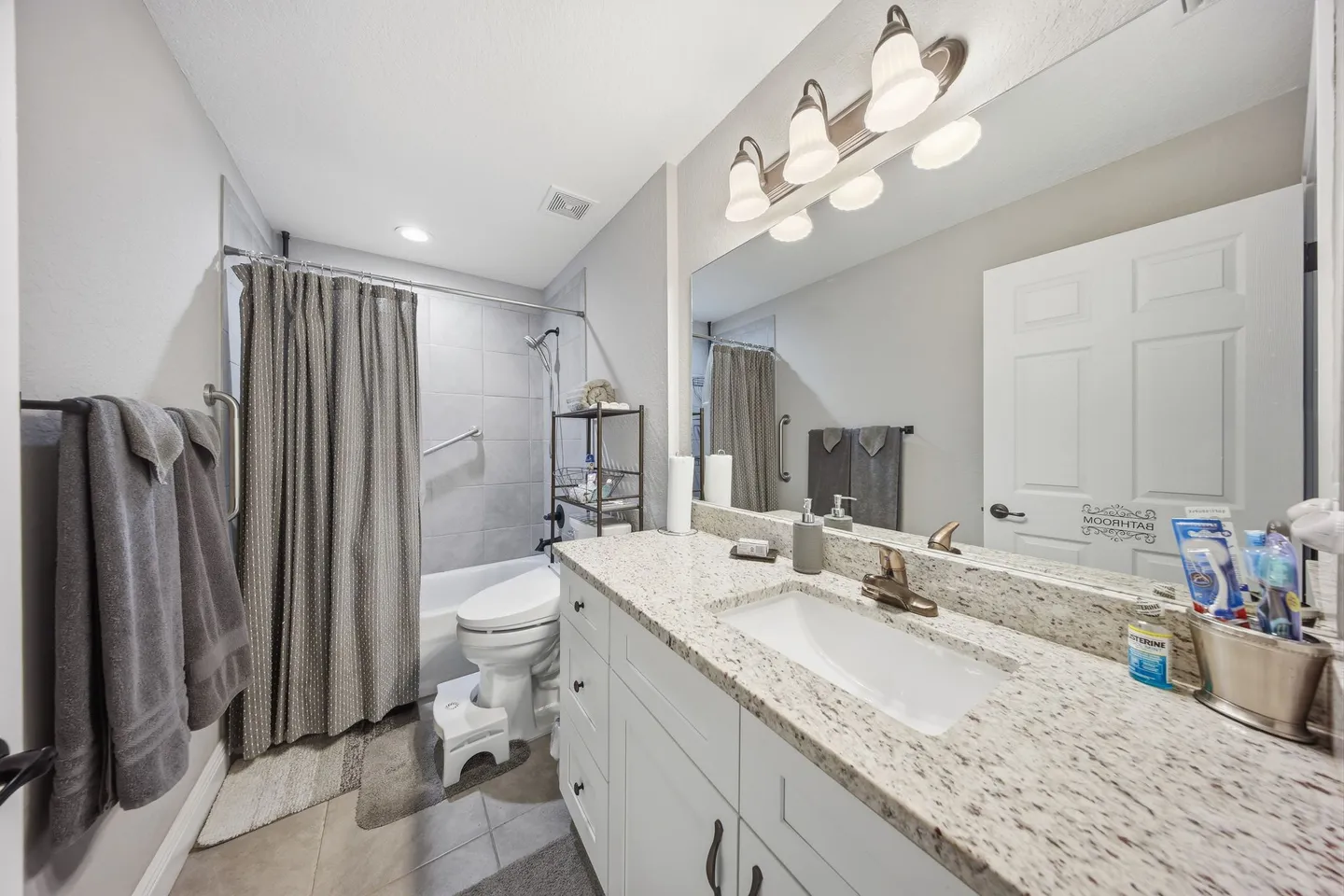 Bathroom with a white vanity, speckled countertop, large mirror with vanity lighting, toilet, and a shower-tub combination with a gray curtain.
