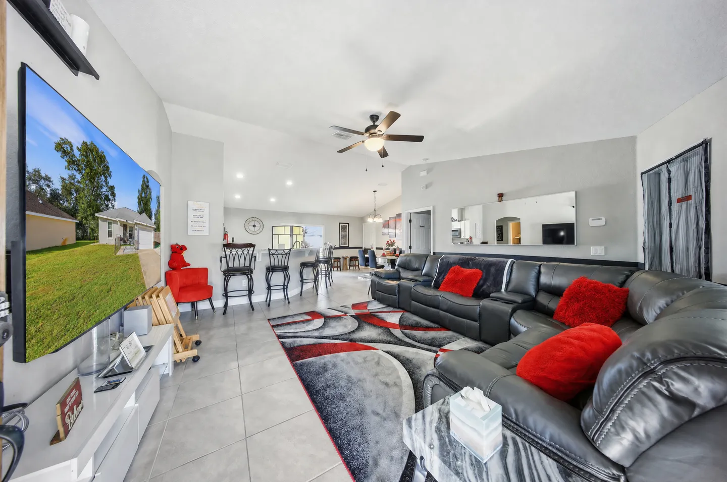 Open-concept living room with a large black sectional sofa, wall-mounted television, tiled flooring, and views into the adjacent kitchen.