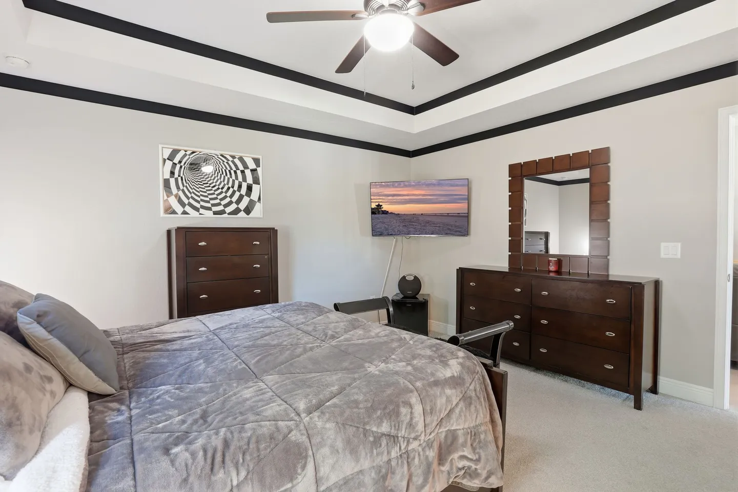 Bedroom featuring a made bed with a plush quilt, two dark wood dressers, a wall-mounted TV, and a tray ceiling with a ceiling fan.
