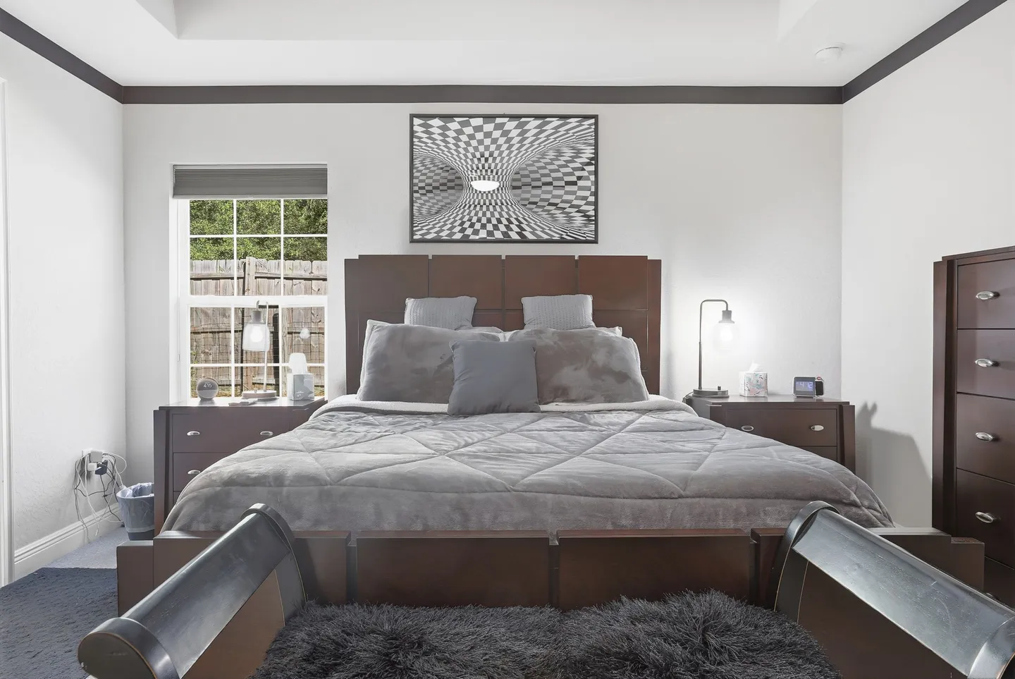 Bedroom featuring a dark wood bed with grey bedding, matching nightstands, a tall dresser, and an optical illusion framed artwork above the headboard.