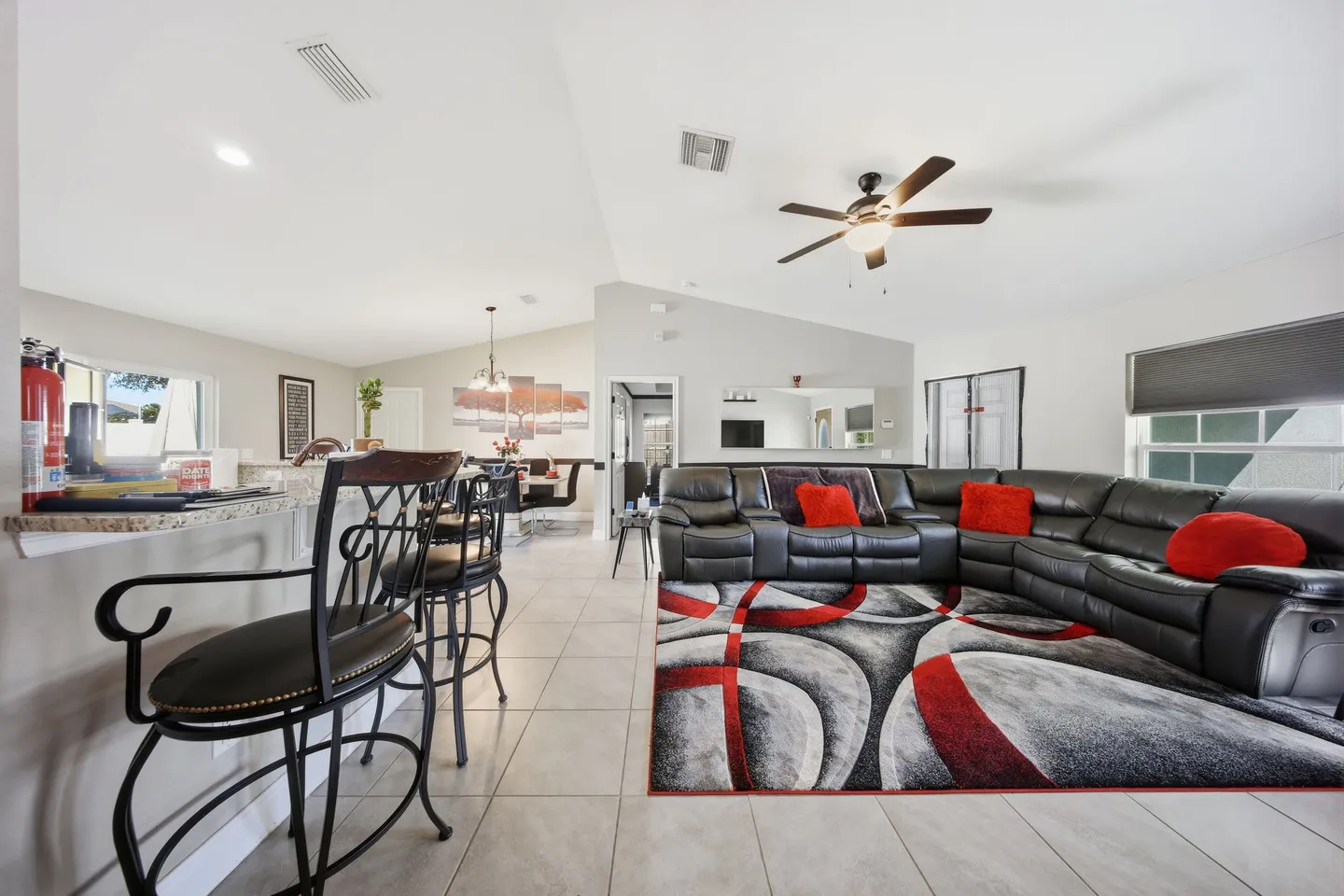 Open-concept living room with a black sectional sofa, patterned area rug, ceiling fan, and breakfast bar with stools.