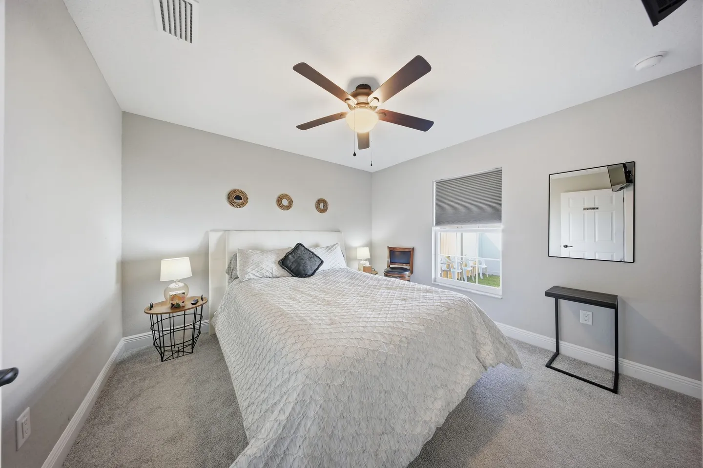 Bedroom with a made bed, carpeted flooring, a ceiling fan, a window, and a wall mirror.