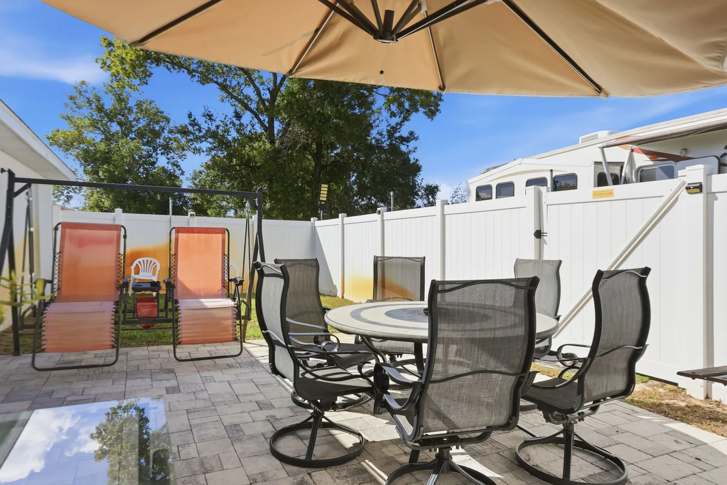 A paved patio with an outdoor dining set under an umbrella, a two-seat swing, and white privacy fencing.