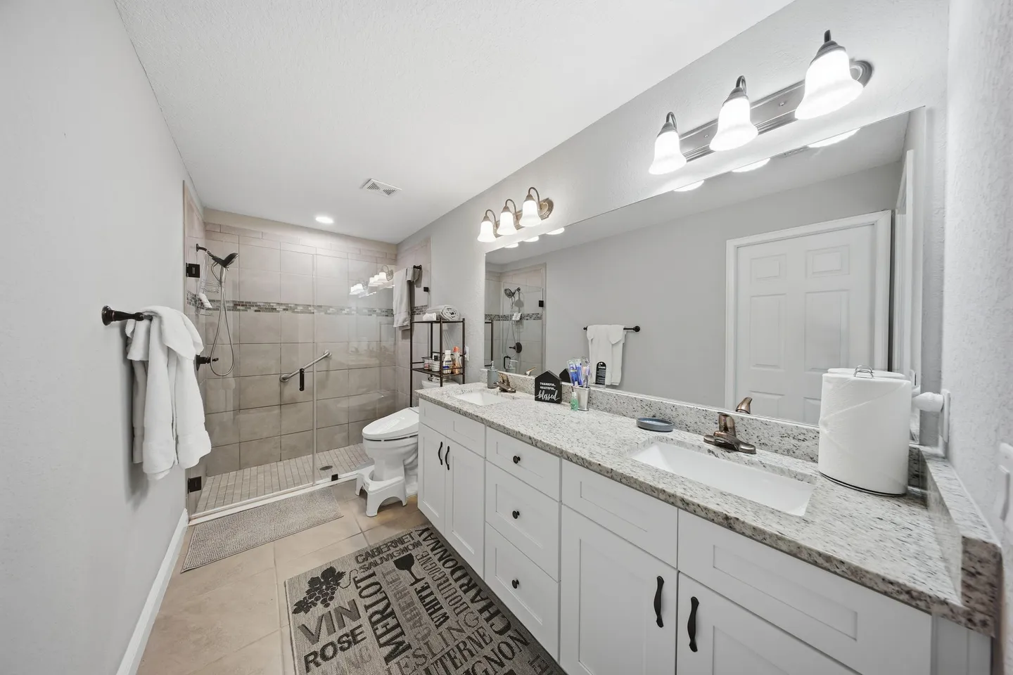 Bathroom with a double-sink white vanity, large mirror with vanity lights, toilet, and a walk-in tiled shower.