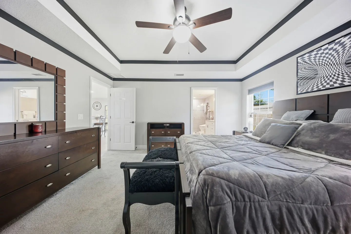 Bedroom with a tray ceiling, ceiling fan, dark wood furniture, a made bed with gray bedding, and doorways leading to a hall and an ensuite bathroom.