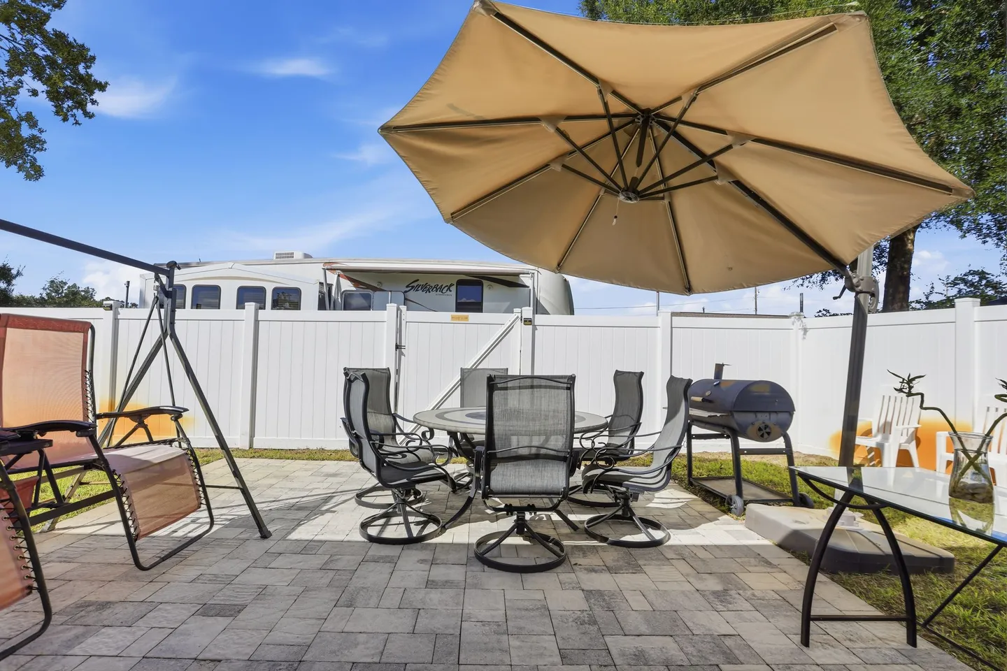Outdoor paved patio area featuring a dining table with chairs under an umbrella, a swing chair, and a barbecue grill, enclosed by a white fence.