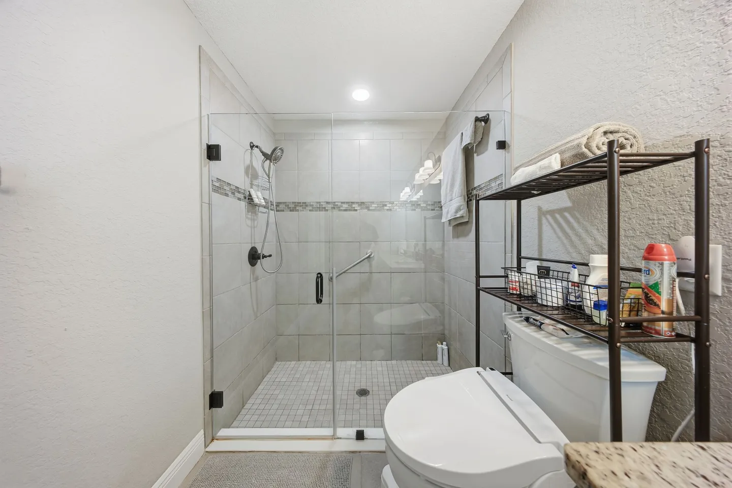 Bathroom showing a walk-in shower with glass doors, a toilet with bidet attachment, and an over-the-toilet shelving unit.