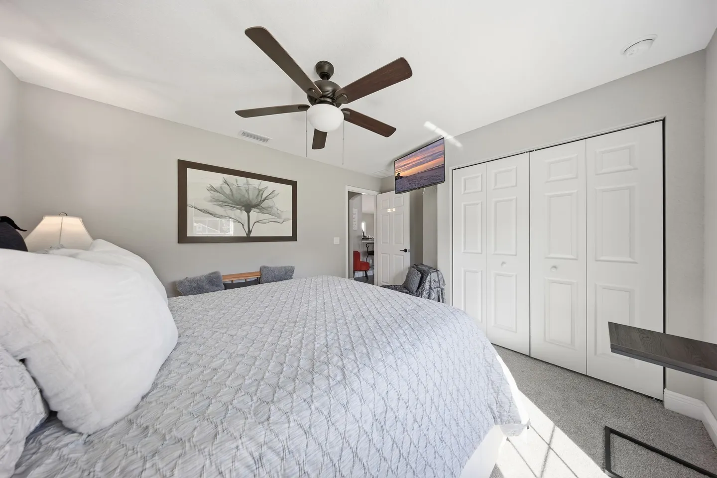 Bedroom with a bed dressed in a gray quilt, ceiling fan, wall-mounted TV, and white closet doors.