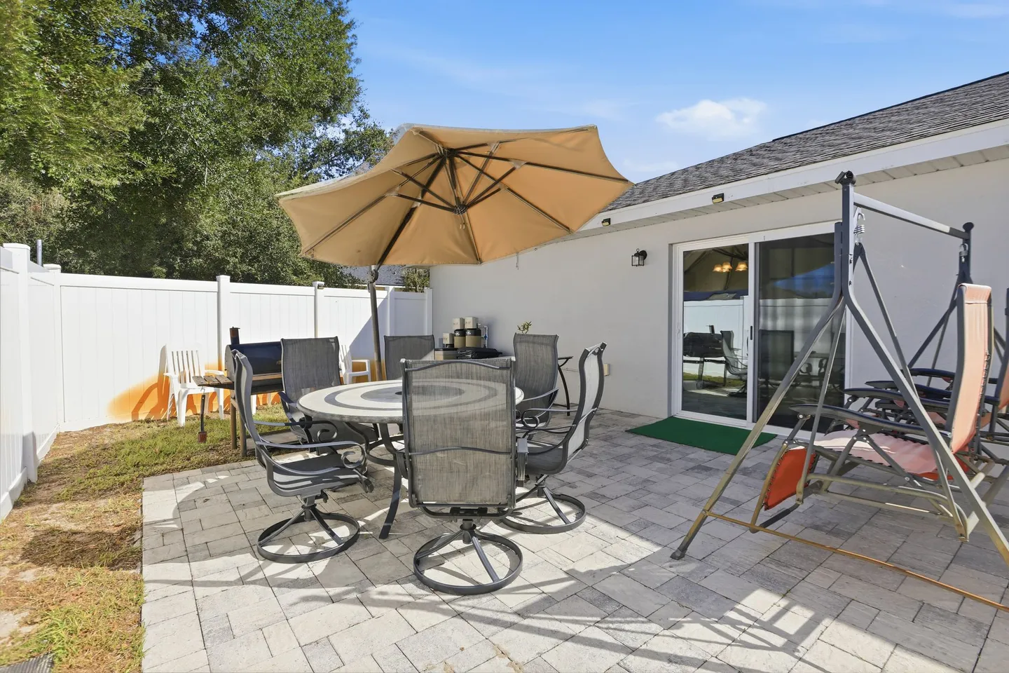 A paved backyard patio featuring an outdoor dining table with chairs under an umbrella, a swing chair, and a barbecue grill next to a white fence.