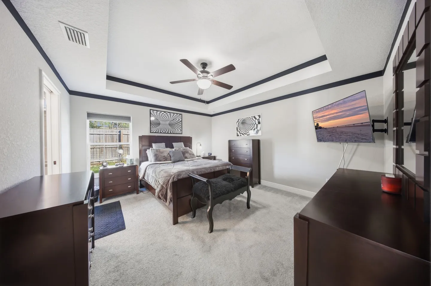 Bedroom with tray ceiling, ceiling fan, dark wood furniture, mounted television, and carpeted floor.