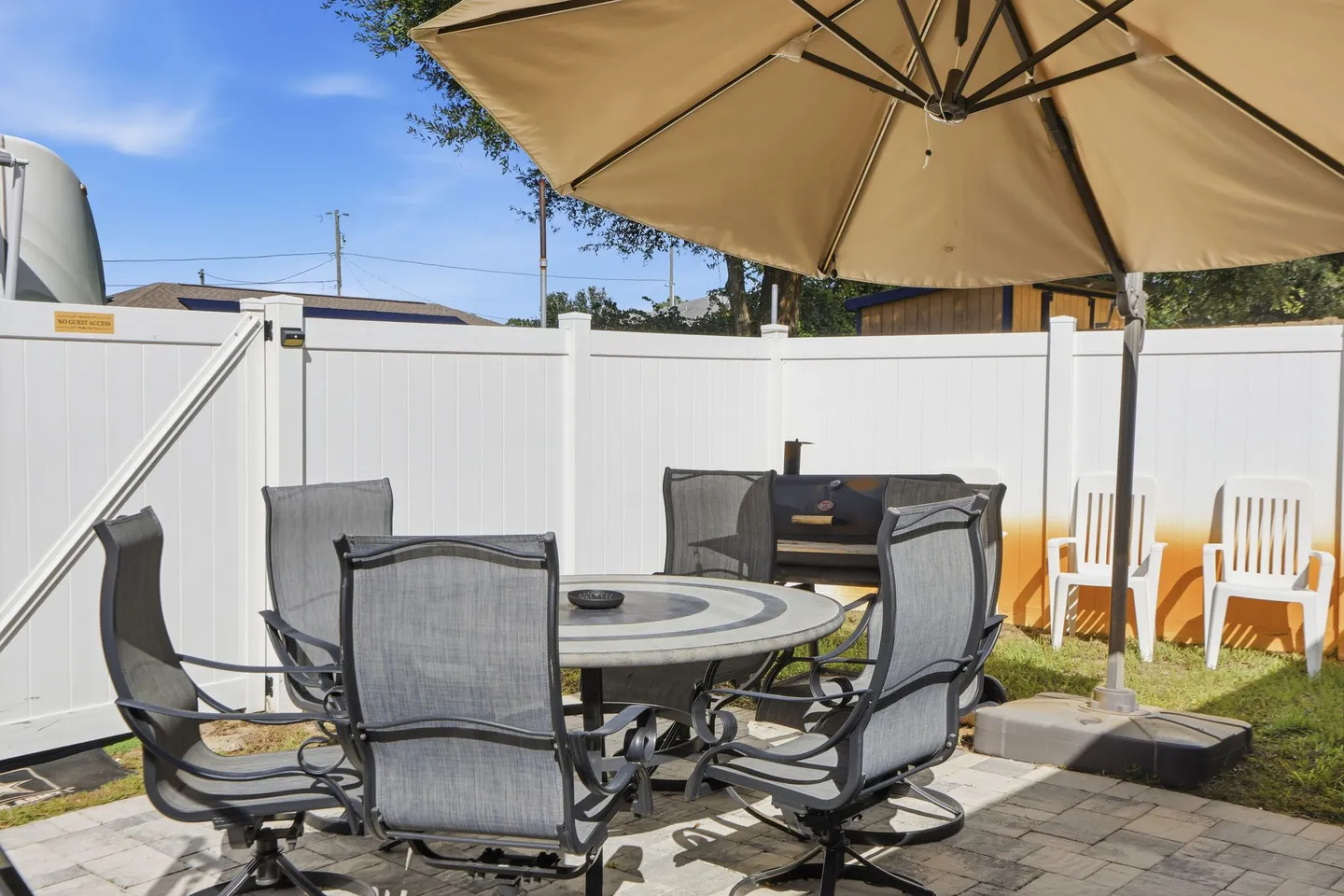 Outdoor patio dining table and chairs shaded by a large umbrella within a fenced yard with a barbecue grill.