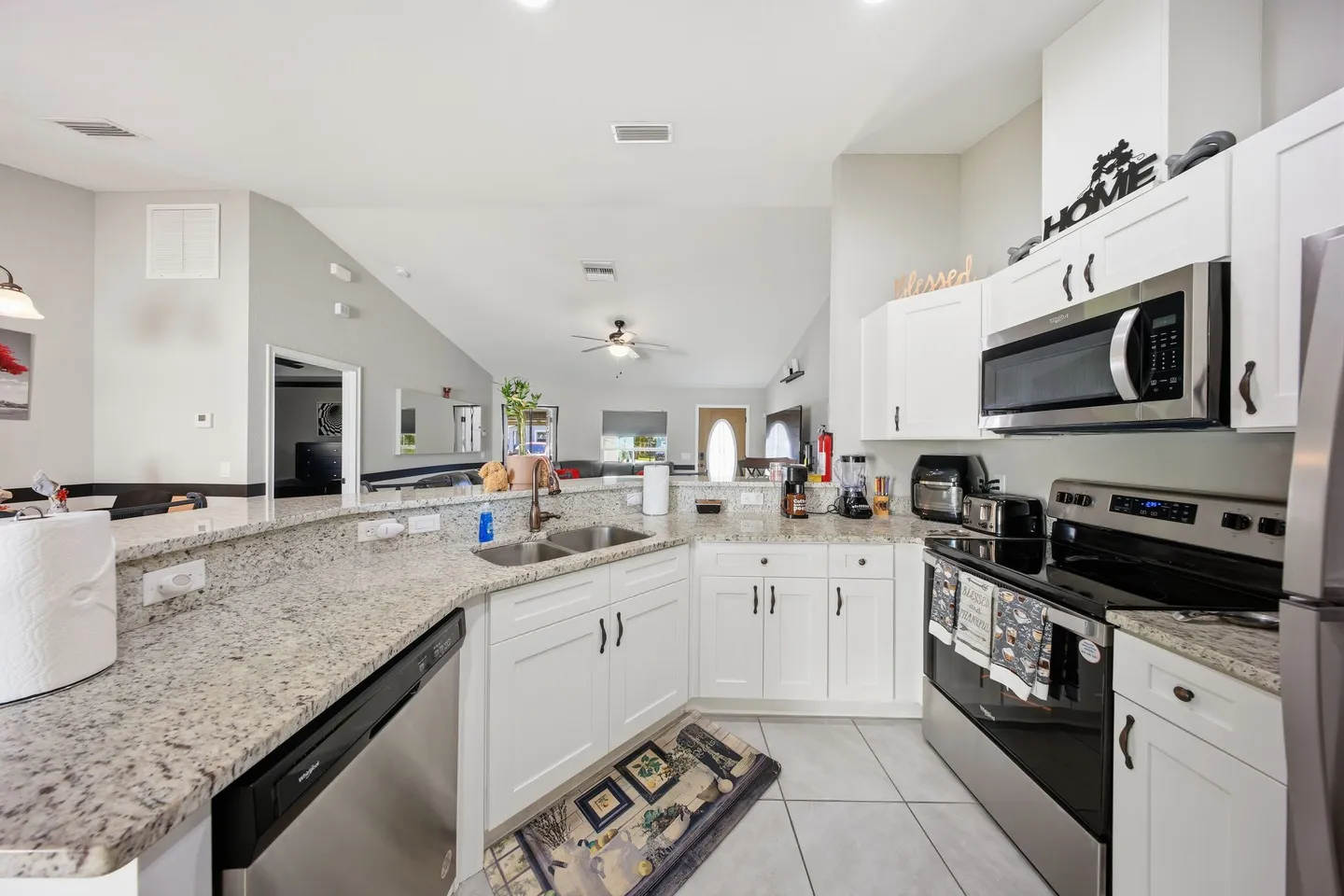 A kitchen featuring white cabinets, speckled granite countertops, stainless steel appliances, and a pass-through bar looking into the living room.