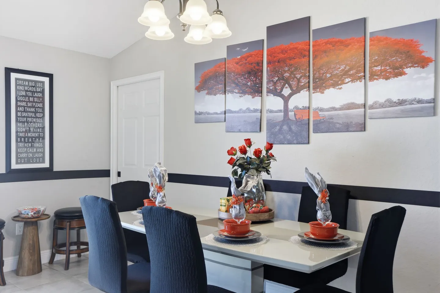 Dining area featuring a modern table set with orange bowls, black upholstered chairs, and a four-panel canvas art piece of an orange tree.