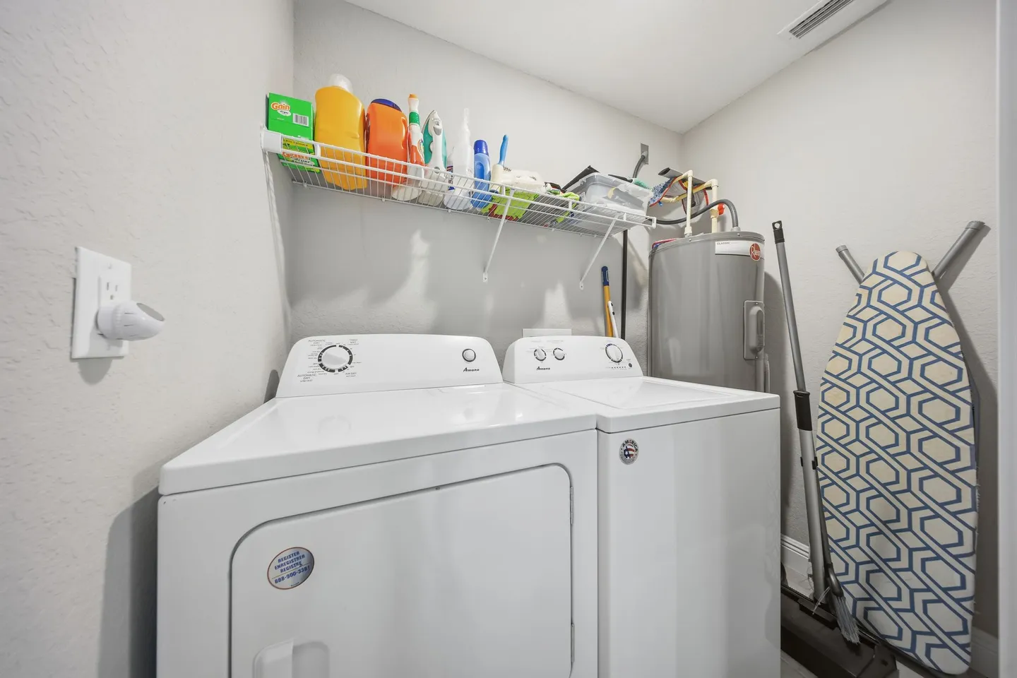 Laundry closet with a white front-load dryer, top-load washer, water heater, ironing board, and wire shelf holding cleaning supplies.