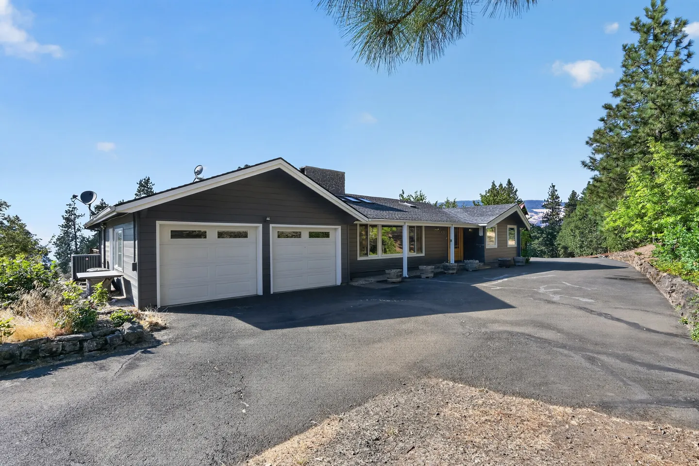 Exterior front view of a single-story dark gray house with a two-car garage and a wide paved driveway under a blue sky.
