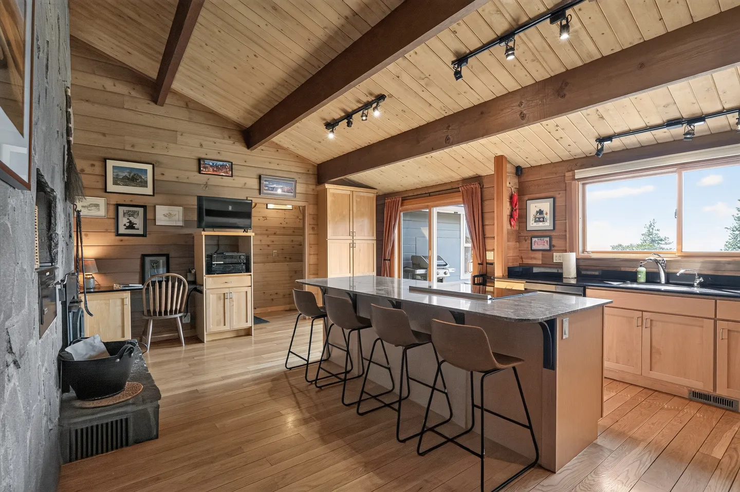 Open-concept kitchen and living area featuring wood-paneled walls and ceiling, a large kitchen island with barstools, and a stone fireplace.