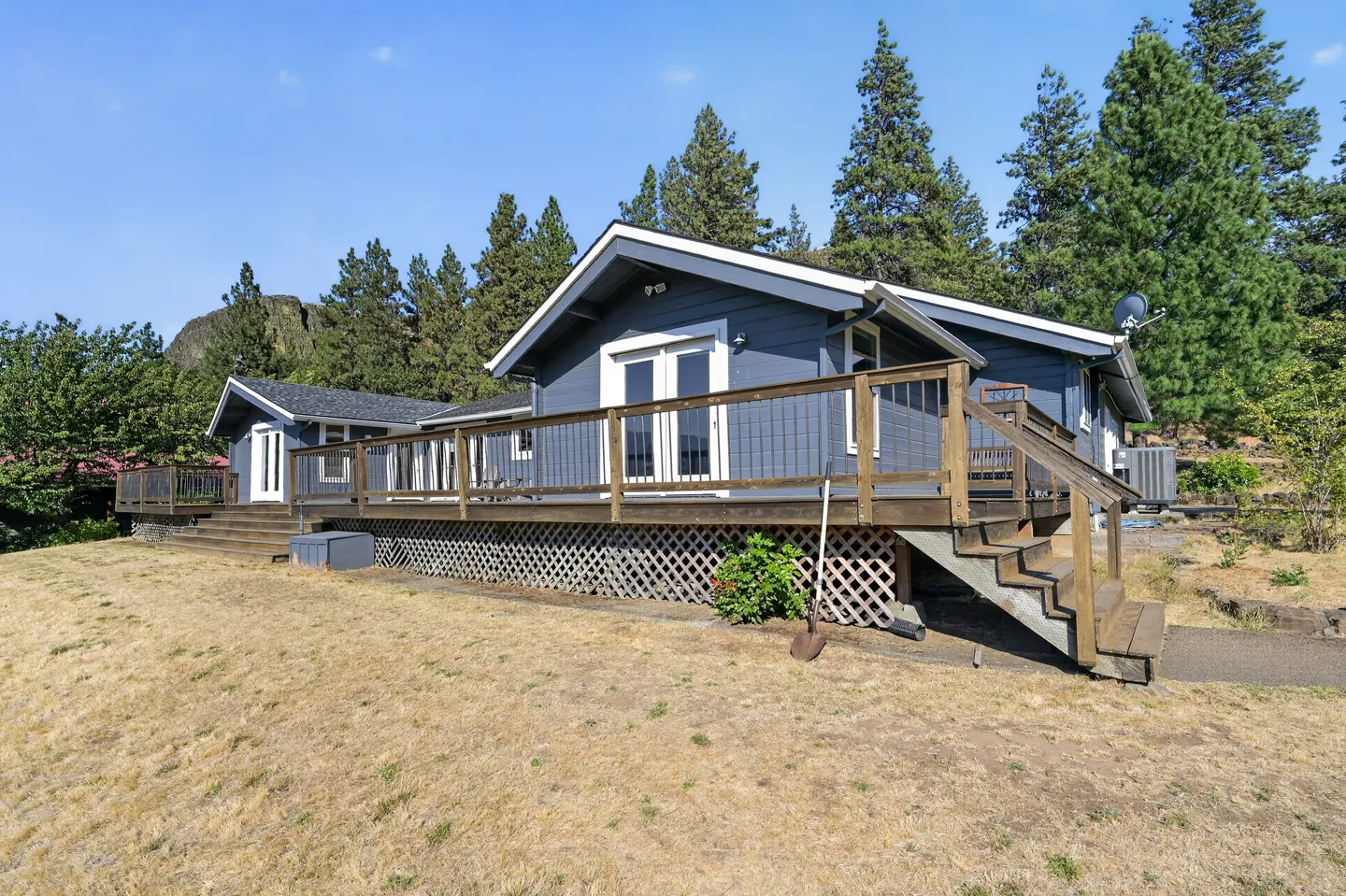 Exterior view of a blue home with a large wooden deck, double French doors, and pine trees under a clear sky.