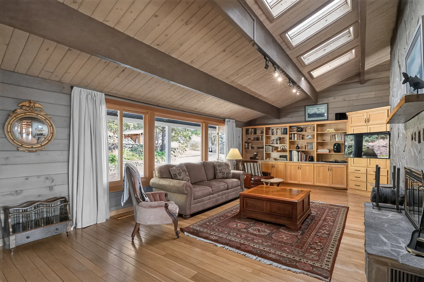 Living room with sloped wood-paneled ceiling, skylights, wood flooring, a sofa, coffee table over an area rug, built-in bookshelves, and large windows.